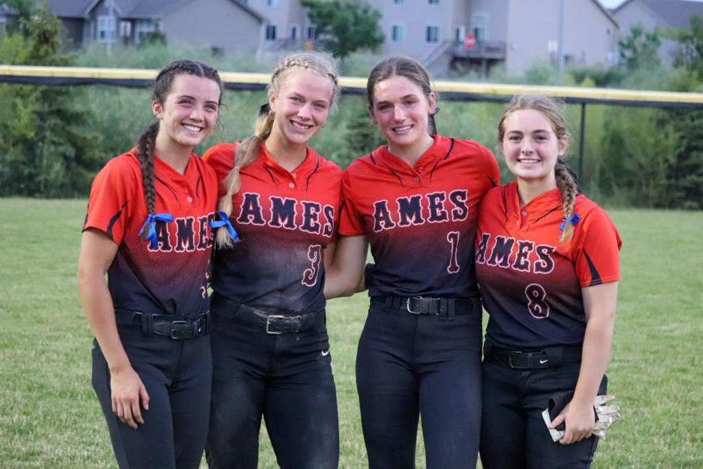 Four female athletes are smiling and wearing softball uniforrms